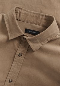 Tan corduroy overhemd met een gevouwen kraag, knoopsluiting met donkere knopen en een zwart label met de tekst "Matinique" aan de binnenkant van de kraag.