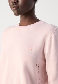 Lys rosa genser laget av mykt materiale med rund hals, med liten brodert logo og tre dekorative stikkmønstre på siden.