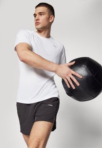 Camiseta deportiva blanca ajustada de manga corta, combinada con pantalones cortos gris oscuro con aberturas laterales. Sosteniendo una pelota medicinal negra.