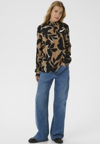 Vrouw in een bruine en zwarte bloemen lange mouw blouse, losse blauwe jeans en zwarte schoenen, staande met één hand in de zak tegen een witte achtergrond.