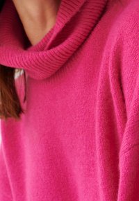 Sweter z dzianiny w kolorze fuksji z szerokim golfem. Miękka tekstura i prążkowane wykończenie wokół kołnierza. Zbliżenie na materiał.