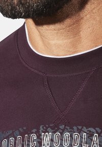Maroon sweatshirt featuring a crew neck and visible stitching. "NORDIC WOODLAND" graphic on front with textured background pattern.