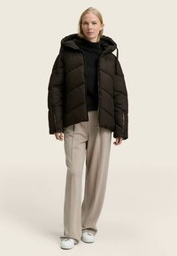 Giacca imbottita nera con cappuccio, caratterizzata da un motivo a chevron e tasche con cerniera, abbinata a pantaloni beige chiaro e sneaker bianche.