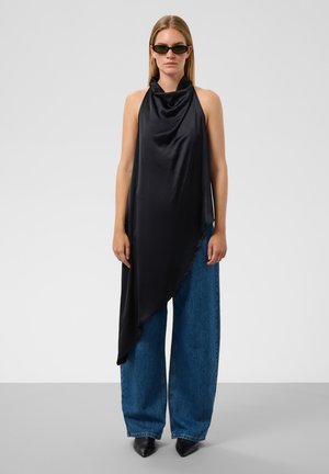 Femme portant un top asymétrique noir sans manches, un jean bleu à jambes larges, des chaussures noires pointues et des lunettes de soleil teintées vertes, debout sur fond blanc.