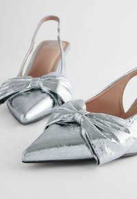 Metallisen hopean väriset slingback-kengät, joissa on terävä kärki ja näyttävä muotoilu edessä. Tasainen pinta ja kiiltävä viimeistely.