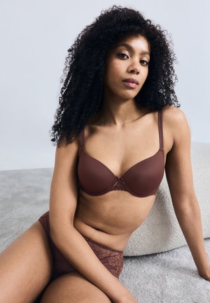 Brown lingerie set featuring a smooth cup bra with lace accents and matching panties. Model poses seated on a soft surface.