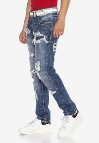 Zerrissene blaue Jeans aus Denim mit mehreren verwaschenen Stellen, schlanker Schnitt und einem kontrastierenden weißen Nieten-Gürtel. Kombiniert mit weißen Sneakers.