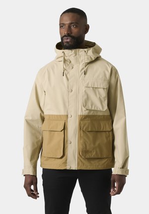 Mężczyzna w beżowo-brązowej kurtce z kapturem i dużymi przednimi kieszeniami, stojący na jednolitym jasnym tle.