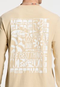 Beige hosszú ujjú póló, fehér grafikai szöveggel és mintákkal, virágos és geometriai elemekkel, puha pamut textúrával.