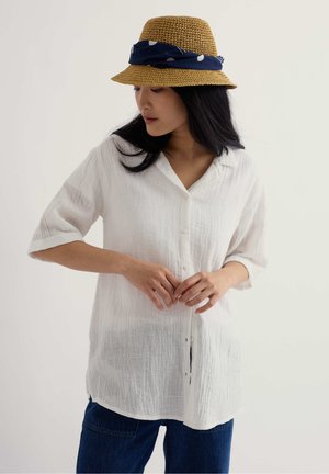 Žena v bielej textúrovanej košeli na gombíky, modrých džínsoch a béžovom pletenom klobúku s námorníckym modrým šálom, ktorá sa pozerá dole doľava.