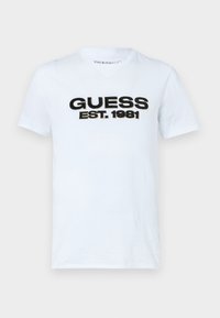 Tricou alb din bumbac cu grafica text "GUESS EST. 1981" în culori negre și aurii pe față. Damage cu mâneci scurte și guler rotund.