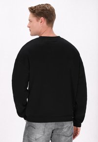 Sort crewneck sweatshirt med sænkede skuldre, struktureret stof og ribbede cuff. Båret med grå jeans, hvilket viser en afslappet pasform.