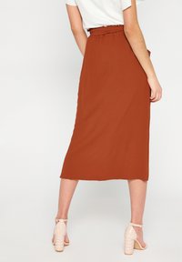Gonna midi color ruggine realizzata con tessuto leggero, con dettaglio in vita e silhouette dritta, abbinata a sandali leggeri con cinturini.