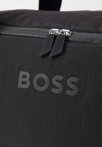 Juodas audinio krepšys su tekstūruota paviršiumi, su užtrauktuku ir guminėmis BOSS logotipu. Įtraukti sidabriniai užtrauktuko priedai ir detalūs akcentai.