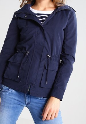 Femme portant une veste zippée bleu marine avec cordons et poches à rabats sur une chemise rayée et un jean bleu clair.