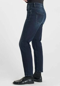 Jean en denim bleu foncé avec une coupe slim, doté d'un design classique à cinq poches et de coutures discrètes. Assortis à des mocassins noirs et des chaussettes grises.