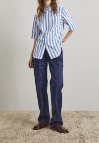 Persona che indossa una camicia a righe verticali blu e bianca, pantaloni cargo blu scuro e scarpe eleganti marroni, in piedi su un tappeto beige.