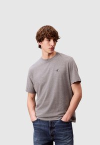 Maglietta di cotone grigia con scollo rotondo e maniche corte; presenta un piccolo logo nero sul petto; abbinata a jeans di denim blu.