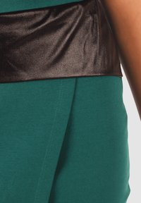 Tejido en color verde azulado con una textura suave, combinado con una cinturilla de satén negro brillante. El diseño presenta pliegues y detalles de costura.