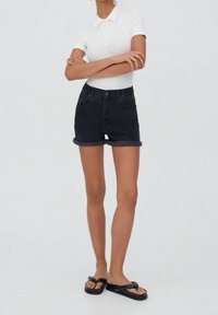 Shorts in denim neri con risvolti, bottone frontale e vita elastica; abbinati a una polo bianca aderente. Le ciabatte sono nere con una fascia.
