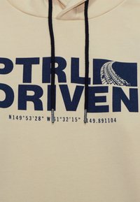 Felpa beige con stampa blu navy che recita "CTRL DRIVEN" e le coordinate. Presenta un cappuccio con coulisse e un tessuto liscio e strutturato.