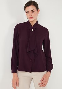 Blusa borgogna con colletto alto, dettaglio in tessuto annodato e bottone circolare al collo. Maniche lunghe con accenti a bottone ai polsini.