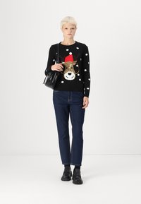 Un pull noir à pois avec un motif de chien portant un chapeau rouge, associé à un jean bleu foncé et des chaussures noires, portant un sac à main noir.