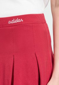 Červená plisovaná sportovní sukně s texturovanou tkaninou a vyšitým logem "adidas" na pasu.