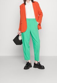 Vrouw in een oranje blazer, witte shirt, groene high-waisted broek, zwarte platform schoenen, met een zwart handtas.