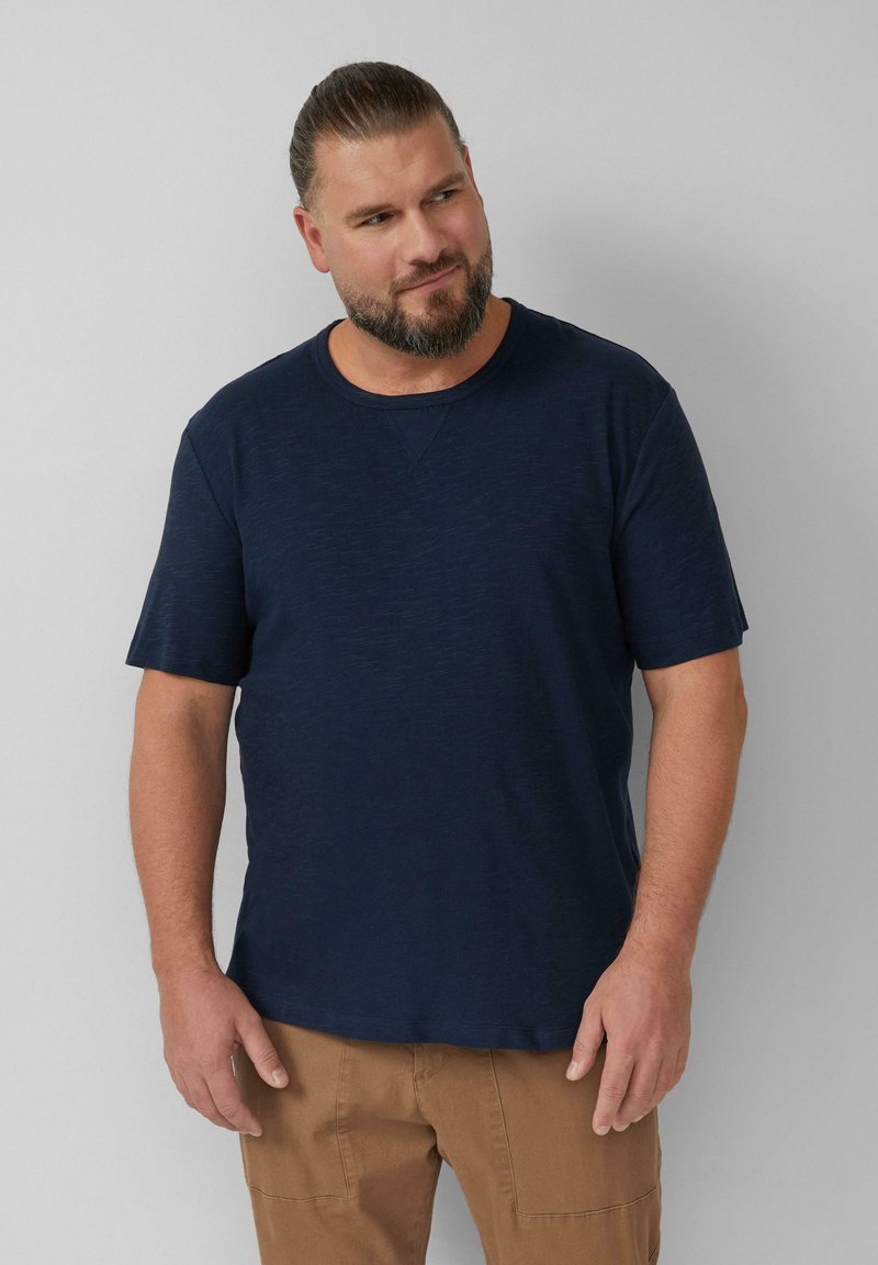 Behaarde man met samengebonden haar, gekleed in een marineblauw overhemd met korte mouwen en een paarse broek, staande tegen een effen lichtgrijze achtergrond.