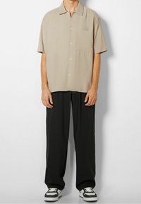 Short-sleeve beige shirt with a chest pocket, paired with loose black pants and black and white sneakers. Smooth texture, structured fit.