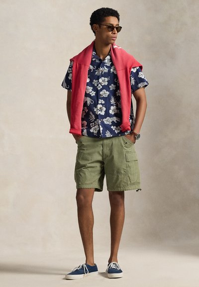 Koszula w kwiaty z krótkim rękawem w kolorach granatowym i białym, zielone szorty cargo z bocznymi kieszeniami, czerwony sweter zarzucony na ramiona i okulary przeciwsłoneczne.