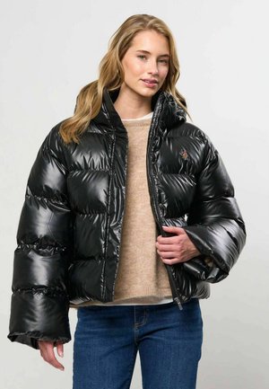 Femme portant une doudoune noire brillante par-dessus un pull beige et un jean bleu, tenant la fermeture éclair de la veste d'une main.