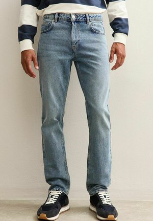 Jeans in denim azzurro chiaro con taglio dritto, design a cinque tasche e chiusura con bottone. Indossati con sneakers nere dotate di lacci bianchi.