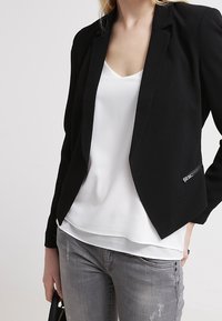 Blazer noir à la coupe structurée, col à revers cranés, et détail de poche zippée, associé à un haut blanc sur un jean gris déchiré.