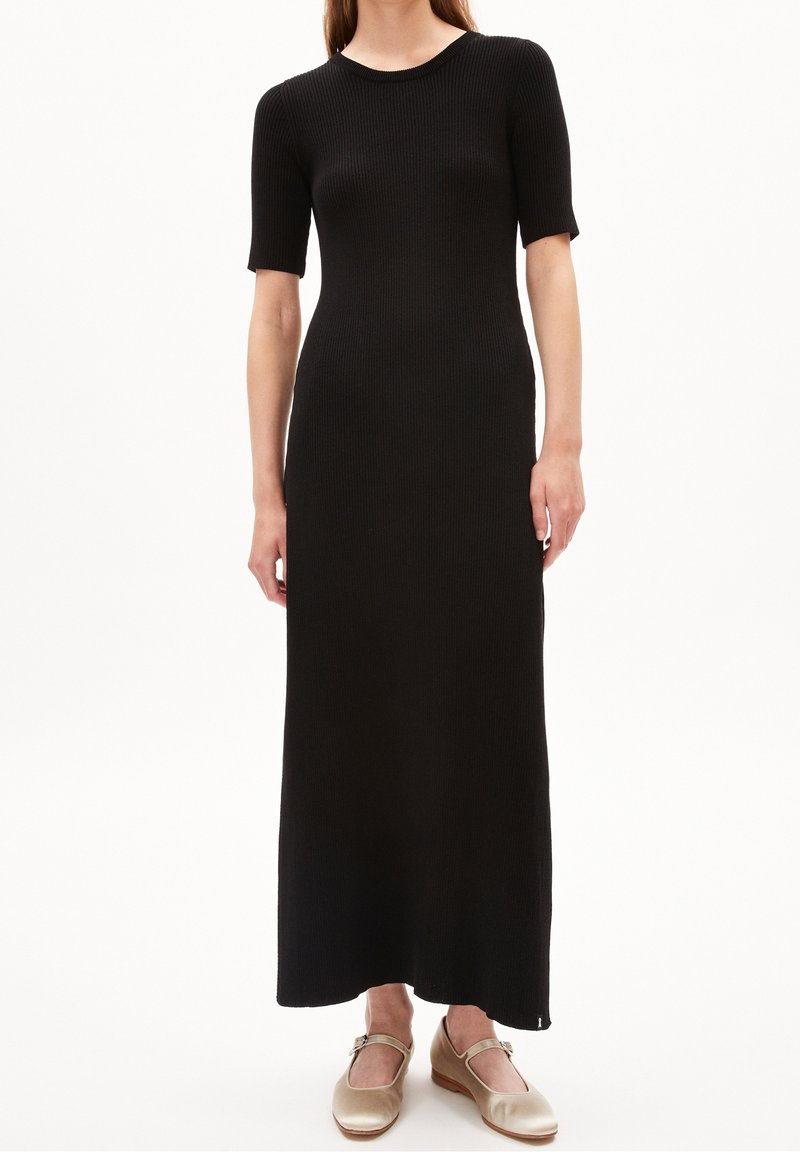 Femme portant une robe longue noire côtelée à manches courtes et des chaussures Mary Jane beiges, debout devant un fond blanc.