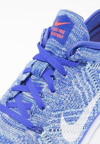 Zapatilla deportiva de punto azul con un upper texturizado, cordones morados, el swoosh blanco de Nike y un logo naranja en la lengüeta. Diseño ligero.