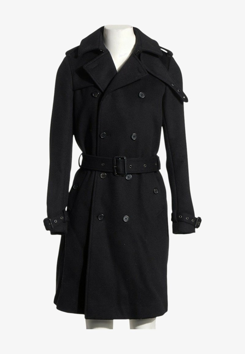 Trench-coat noir croisé à larges revers, ceinturé à la taille et avec des sangles de poignet réglables sur un mannequin.