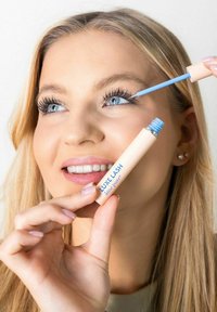 Ljust rosa mascararör med blå text, hålls i en hand; en blå applikatorstav sträcker sig mot långa ögonfransar. Klar, ljus bakgrund.