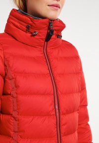Veste matelassée rouge avec un col montant, un matelassage texturé, une fermeture éclair à l'avant et des bordures contrastantes. Dispose de cordons de serrage au niveau du col.