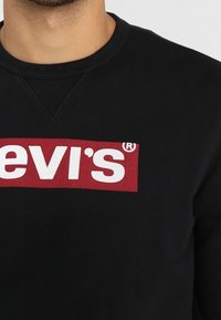 Homme portant un sweat-shirt noir à col rond avec un patch logo rectangulaire rouge affichant le texte blanc « Levi's » sur la poitrine.