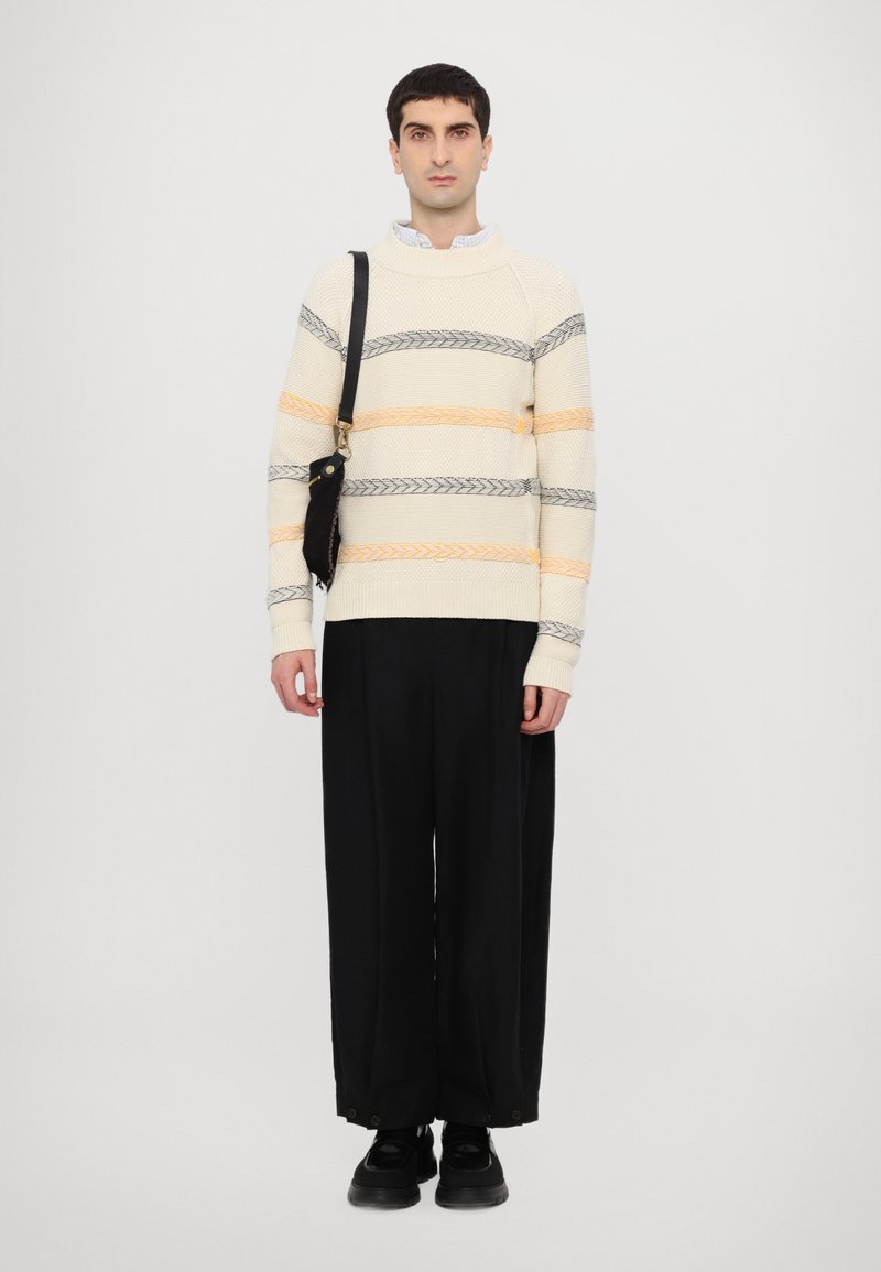 Pull en tricot crème avec des rayures grises et orange, associé à un pantalon noir ample et des chaussures noires. Le modèle porte un sac à bandoulière noir.