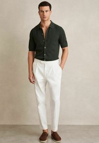 Cămașă tricotată cu mânecă scurtă, de culoare verde închis, cu guler, purtată cu pantaloni albi plisați și pantofi slip-on din nubuc maro.