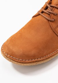 Chaussure en daim marron avec un bout rond, couture beige le long de la semelle et lacets marron assortis sur un fond blanc.