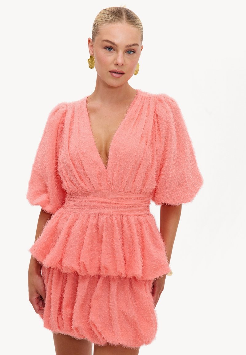 Femme aux cheveux blonds plaqués en arrière, portant de grandes boucles d'oreilles en or et une robe rose duveteuse avec des manches bouffantes et une jupe froncée.