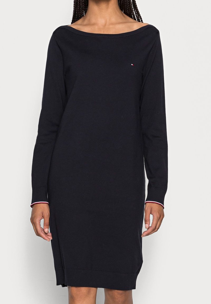 Femme portant une robe noire à manches longues, longueur genou, avec un large décolleté et un petit logo sur la poitrine.