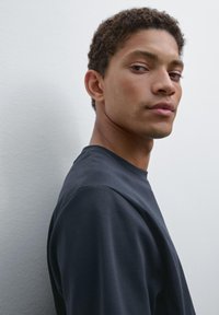 Giovane uomo con capelli ricci corti che indossa una camicia scura, mentre guarda sopra la spalla contro uno sfondo chiaro e semplice.