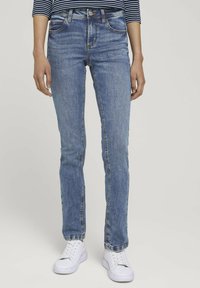 Jean en denim bleu avec une coupe slim, dotés d'un design classique à cinq poches, de légères décolorations et de détails de couture discrets, associés à des baskets blanches.