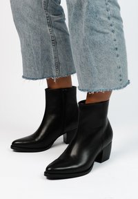 Stivali alla caviglia in pelle nera con punta allungata, tacco squadrato e zip laterale. Indossati con jeans azzurri con orlo sfrangiato.