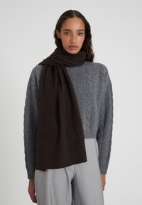 Sciarpa di lana marrone scuro avvolta attorno al collo, abbinata a un maglione grigio chiaro a trecce, che mette in evidenza texture morbide e colori a contrasto.